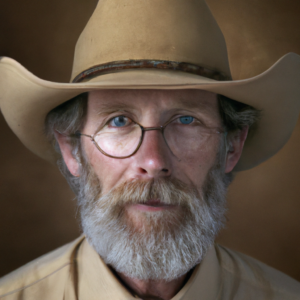 A professional portrait photo of Michael, 62, a small, slim bearded cowboy wearing a crumpled straw cowboy hat, round glasses, blue eyes, work shirt. He\'s looking the camera in the eye.